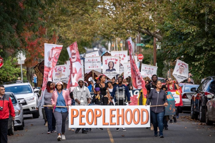 A crowd of people holding banners and signs walking towards the camera on a street lined with parked cars and overhanging leafy branches.