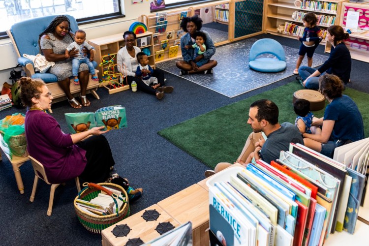 Twelve people sitting in a circle on a carpeted floor looking at a women holding an open children's book.