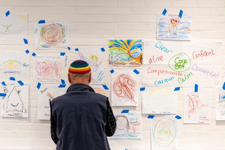 A person wearing a multi-colored kippah looks at a wall of hand-drawn art.